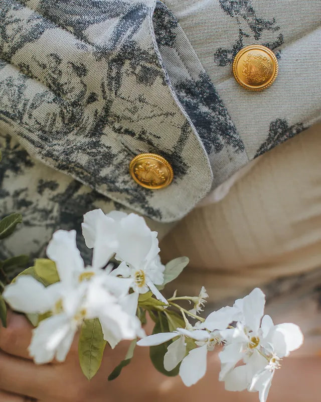 Two gold buttons embossed with the coat of arms on the strap of a MonDoudou Toile de Jouy, a sprig of white blossom in front.