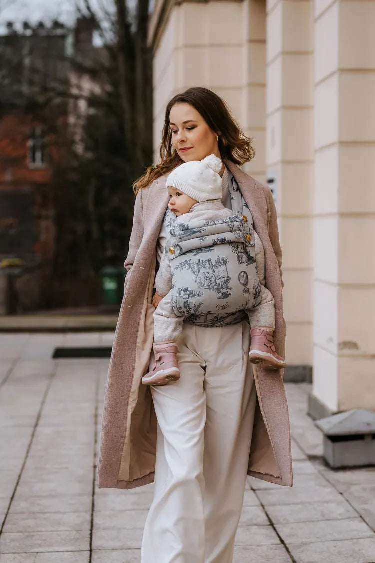 A woman in a wool coat carries her child on her front in the MonDoudou Toile de Jouy.