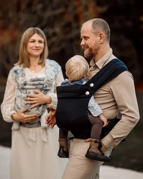 A woman wearing the MonDoudou Toile de Jouy and a man wearing the MonDoudou Noir each carry a child; he turns to her, laughing.