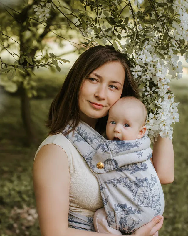 The wearer holds her child in the MonDoudou Toile de Jouy, cheek to cheek, white blossom above them.